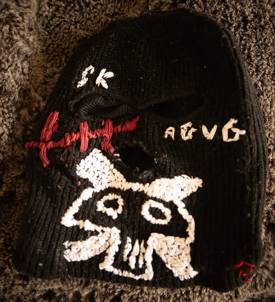 Ski mask Nº 3 laid flat: black knit, large skull embroidered in white thread, SK and AGVG lettering, stitched red cross