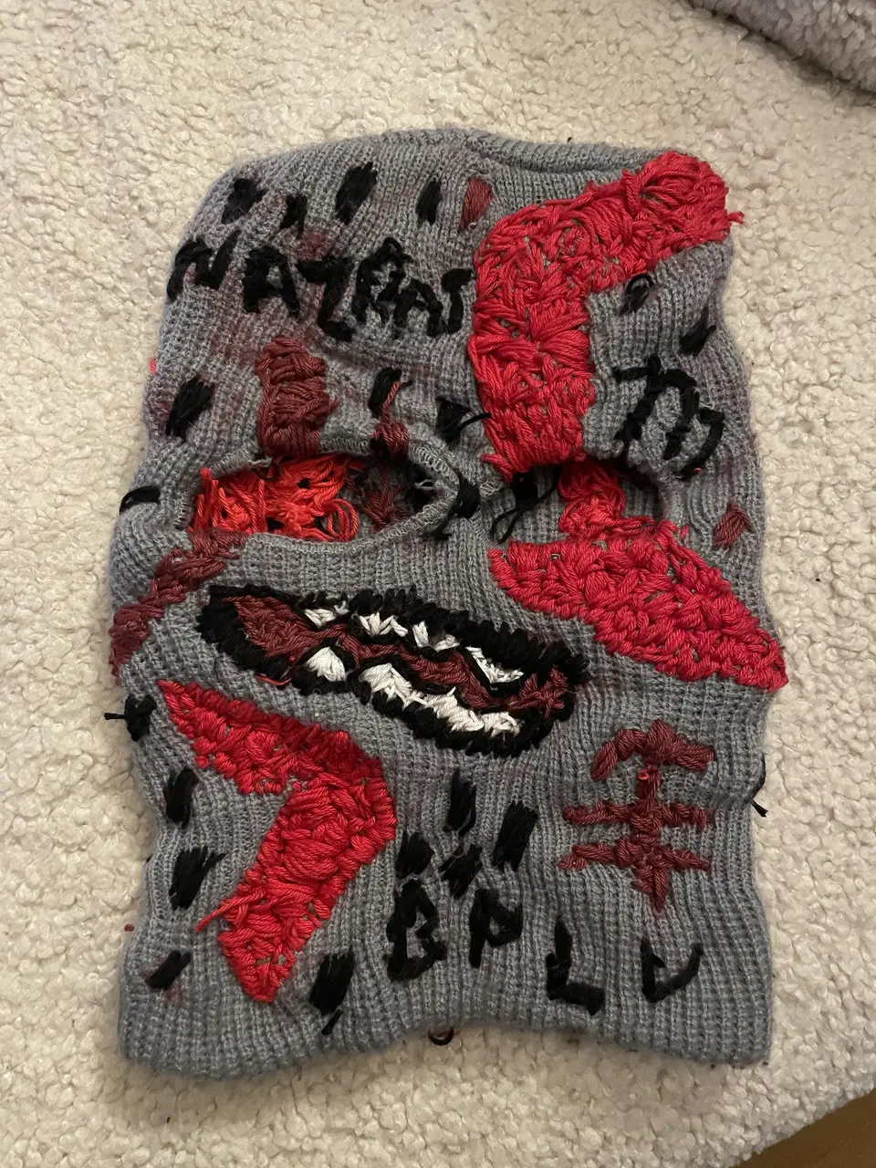 Ski mask Nº 7 laid flat: gray knit, raised red shapes, black hatching, mouth with white teeth, NAZRAT lettering
